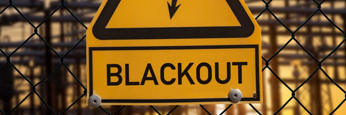 Ein gelbes Warnschild an einem Maschendrahtzaun mit einem schwarzen Blitz-Symbol und der Aufschrift „BLACKOUT“. Im Hintergrund ist unscharf eine elektrische Anlage mit Strommasten zu sehen.