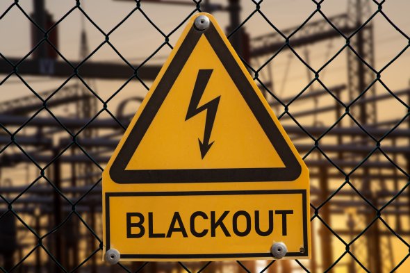 Ein gelbes Warnschild an einem Maschendrahtzaun mit einem schwarzen Blitz-Symbol und der Aufschrift „BLACKOUT“. Im Hintergrund ist unscharf eine elektrische Anlage mit Strommasten zu sehen.