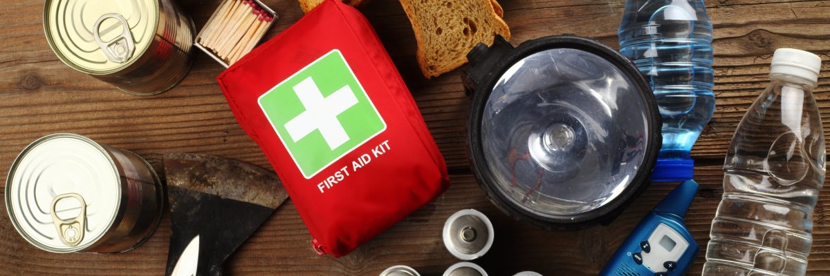 Notfallausrüstung auf Holztisch Auf einem Tisch ist eine Zusammenstellung von Notfallausrüstung zu sehen. Dazu gehören mehrere Konservendosen, Brotstücke, ein rotes Erste-Hilfe-Set, eine große schwarze Taschenlampe mit dazugehörigen Batterien, zwei Plastikwasserflaschen, ein Taschenmesser, eine Axt, weiße Kerzen und eine Schachtel Streichhölzer.
