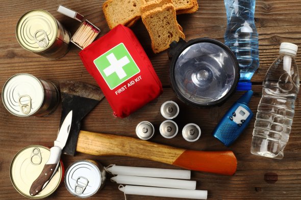 Auf einem Tisch ist eine Zusammenstellung von Notfallausrüstung zu sehen. Dazu gehören mehrere Konservendosen, Brotstücke, ein rotes Erste-Hilfe-Set, eine große schwarze Taschenlampe mit dazugehörigen Batterien, zwei Plastikwasserflaschen, ein Taschenmesser, eine Axt, weiße Kerzen und eine Schachtel Streichhölzer.