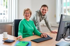 Frau und Mann am Computer Eine ältere Frau in einem grünen Pullover sitzt an einem Computer und lächelt in die Kamera. Ein jüngerer Mann in einem grauen Hemd beugt sich über ihre Schulter und lächelt ebenfalls. Auf dem Schreibtisch befinden sich eine Tastatur, eine Maus, ein blauer Notizblock, ein Taschenrechner, ein Glas Wasser und eine Tasse.