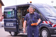 Mitarbeiter der Stadtwerke Overath Ein lächelnder Mann in blauer Arbeitskleidung mit orangefarbenem Gehörschutz und einer Fernbedienung in der Hand steht vor einem blauen Firmenfahrzeug der Stadtwerke Overath.