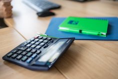 Taschenrechner und Notizbuch Ein blauer Taschenrechner, ein grünes Notizbuch und ein grüner Stift liegen auf einem hellen Holztisch. Im unscharfen Hintergrund sind eine Tastatur und eine Computermaus zu sehen.