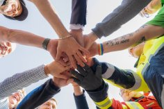 Hände im Kreis Eine Untersicht-Aufnahme einer Gruppe von Personen, die ihre Hände im Zentrum eines Kreises aufeinanderlegen. Die Personen tragen unterschiedliche Kleidung, darunter Sicherheitswesten in Gelb und Orange sowie Arbeitshandschuhe.