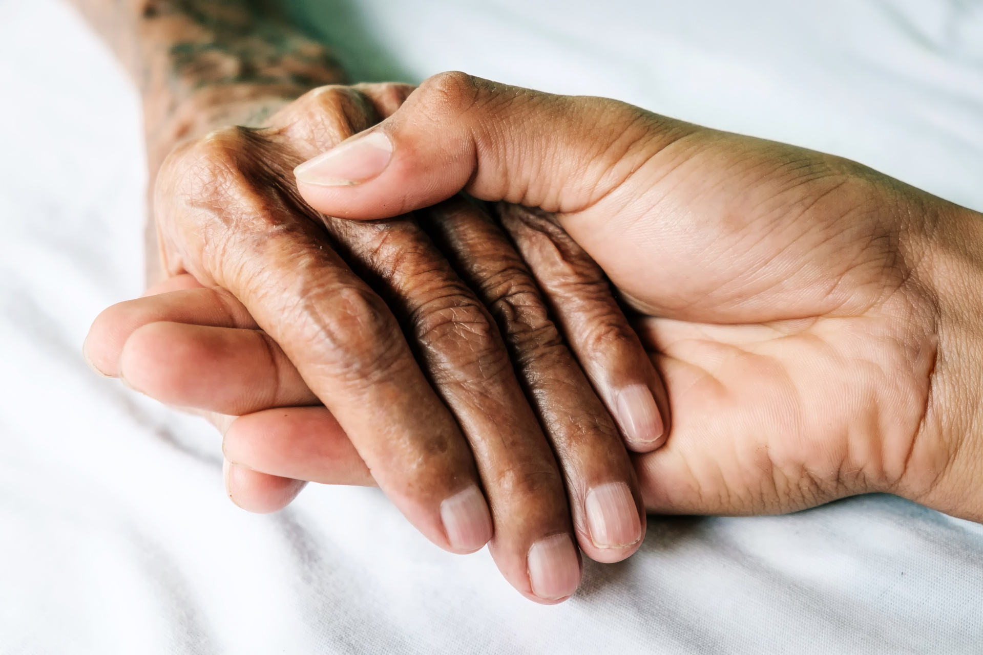 Hand hält ältere Hand Nahaufnahme von zwei Händen auf einem weißen Untergrund. Eine glatte Hand liegt schützend auf einer älteren, faltigen Hand.