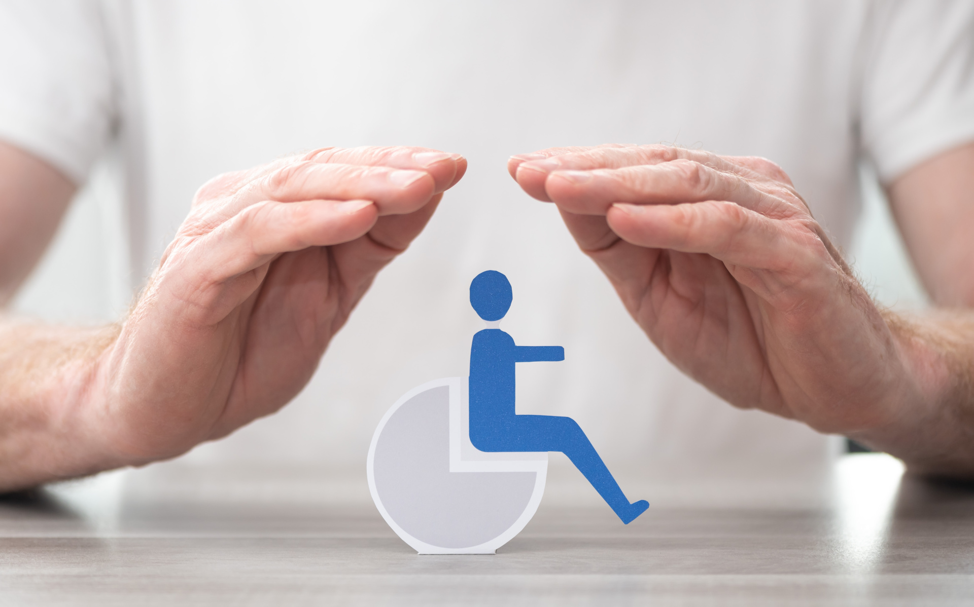 Rollstuhl-Symbol zwischen zwei Händen Eine blaue Papierfigur eines Rollstuhlfahrers steht auf einer grauen Fläche. Zwei Hände sind schützend um das Symbol herum positioniert.