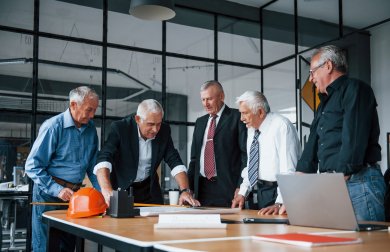 Fünf ältere Männer in Business-Kleidung stehen in einem modernen Büro mit Glaswänden um einen Tisch. Sie blicken gemeinsam auf Baupläne, auf die einer der Männer zeigt. Auf dem Tisch liegen zudem ein orangefarbener Schutzhelm, ein Laptop und ein rotes Notizbuch.