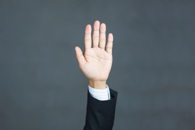 Eine Person in einem dunklen Anzug mit einem weiß-blau gestreiften Hemd hebt die Hand. Die Handfläche ist offen und zeigt nach vorne. Der Hintergrund ist eine schlichte, graue Fläche.