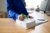 Eine Person in einem blauen Hemd markiert mit einem grünen Textmarker Unterlagen in einem weißen Ordner auf einem Holztisch. Im Hintergrund sind unscharfe Computermonitore zu sehen.