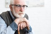 Portrait eines älteren Mannes mit weißem Bart und Brille. Er trägt eine graue Weste über einem hellblauen Hemd und stützt seine Hände auf einen hölzernen Gehstock.