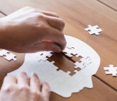 Eine Person setzt auf einem Holztisch ein weißes Puzzle zusammen, dessen Teile die Silhouette eines menschlichen Kopfes bilden. Eine Hand platziert gerade ein Puzzleteil in eine Lücke.