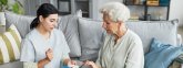 Eine Pflegekraft und eine ältere Frau mit weißen Haaren sitzen gemeinsam auf einem Sofa. Die Pflegekraft hält eine Medikamentenbox in der Hand und erklärt deren Nutzung. Auf einem Couchtisch im Vordergrund liegen ein Inhalator und ein kleines Medikamentengläschen.