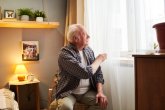Ein älterer Mann mit weißen Haaren sitzt in einem Sessel und blickt durch weiße, transparente Vorhänge aus einem Fenster.