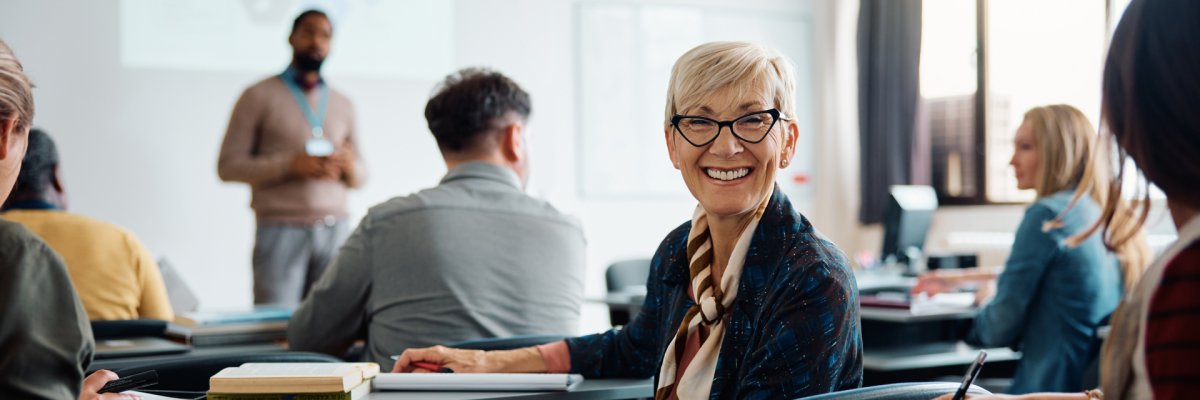 Eine blonde Frau mit Brille lächelt in einem Kursraum der Volkshochschule. Sie sitzt an einem Tisch mit Notizbüchern. Im Hintergrund ist ein Dozent vor einer Leinwand sowie weitere Kursteilnehmer zu sehen.