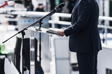 Ein Mann in einem steht an einem transparenten Rednerpult. Ein Mikrofon auf einem Stativ ist vor ihm positioniert.