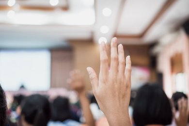 Eine erhobene Hand im Vordergrund, die in einer Gruppe von Menschen in einem hellen Raum nach oben gestreckt ist.