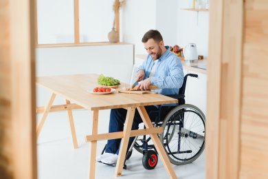 Ein Mann sitzt in einem Rollstuhl an einem Holztisch und schneidet Brot auf einem Holzbrett. Auf dem Tisch befinden sich zudem eine Schale mit Salat und ein Teller mit Kirschtomaten.