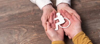 Zwei Paare Hände halten gemeinsam einen weißen Papierschnitt des internationalen Symbols für Barrierefreiheit (eine Person im Rollstuhl).