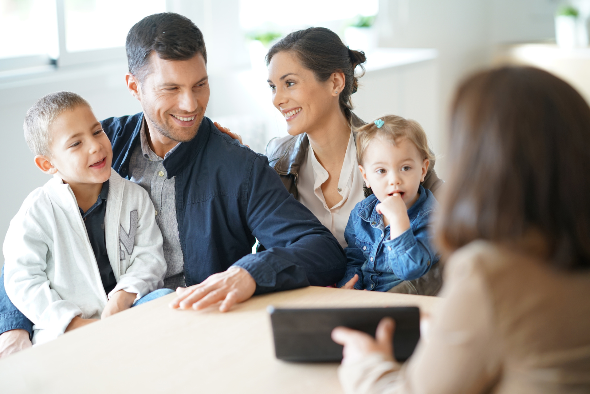 Beratungsgespräch einer Familie Eine vierköpfige Familie – Vater, Mutter, ein Junge und ein kleines Mädchen – sitzt lächelnd an einem hellen Holztisch. Sie blicken eine Person an, die von hinten zu sehen ist und ein schwarzes Tablet hält.