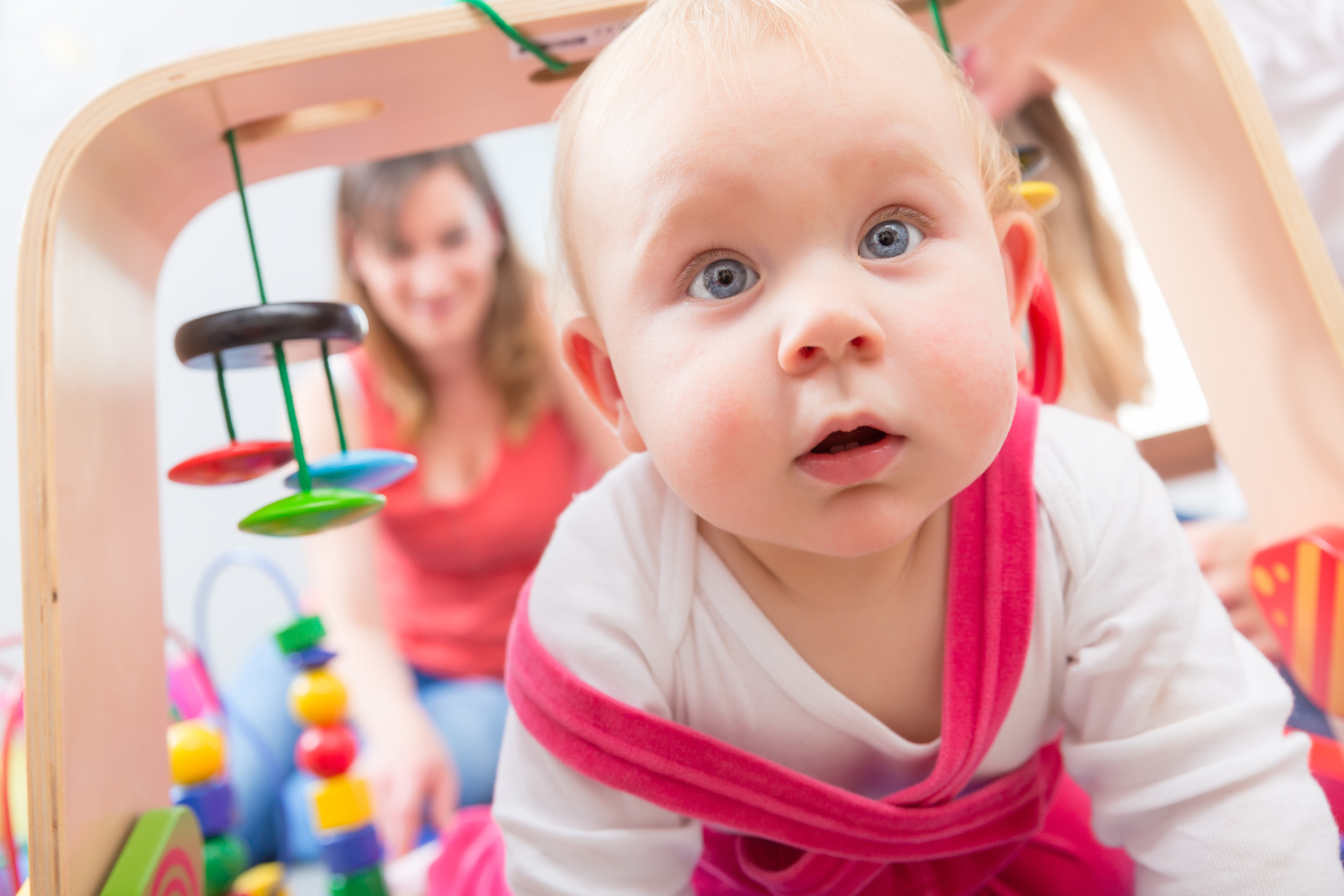 Baby im Spielbogen Ein Baby in rosa-weißer Kleidung blickt unter einem hölzernen Spielbogen mit bunten Elementen in die Kamera.