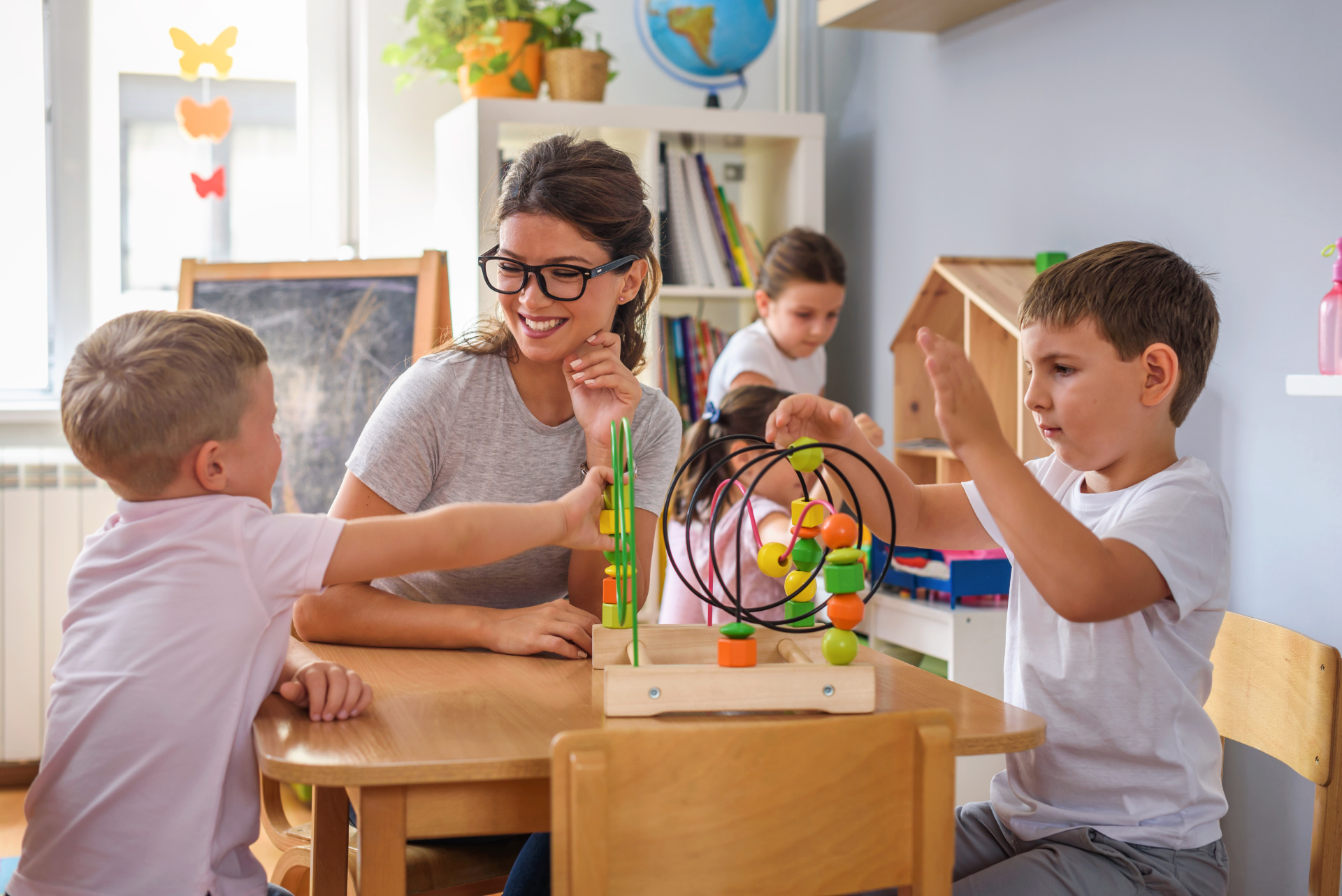 Kinder spielen mit Drahtlabyrinth Zwei Jungen spielen an einem Holztisch mit einem bunten Drahtlabyrinth, während eine lächelnde Frau mit Brille sie beobachtet.