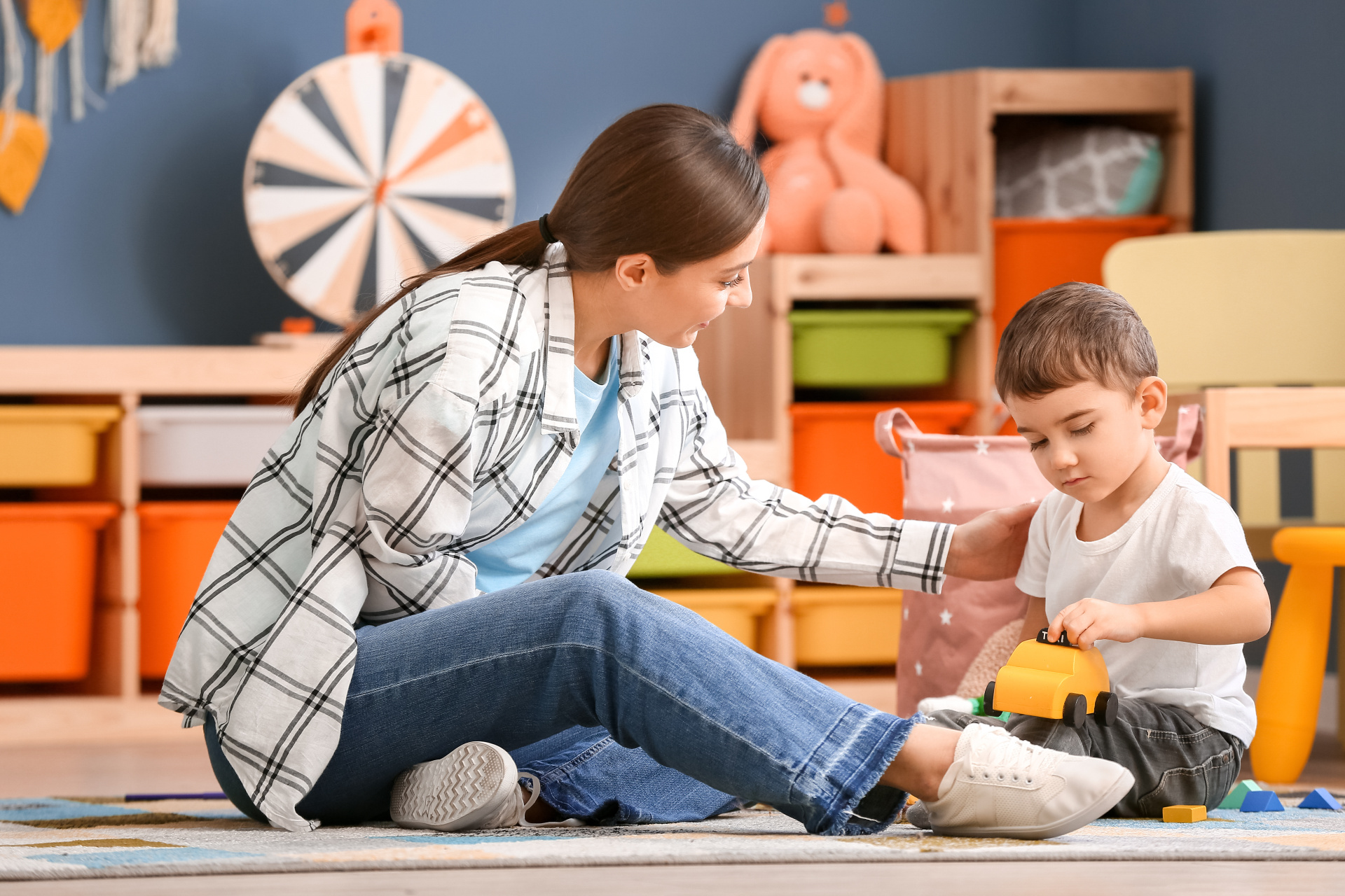 Kind spielt mit Spielzeugauto Eine Erzieherin mit einen kleinen Jungen, der auf dem Boden eines Spielzimmers mit einem gelben Spielzeugauto spielt.