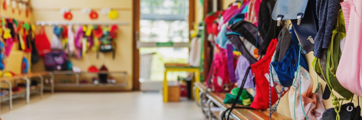 Kindergarderobe Bunte Jacken und Taschen hängen an Haken an einer hellgelben Wand in einem hellen Raum.