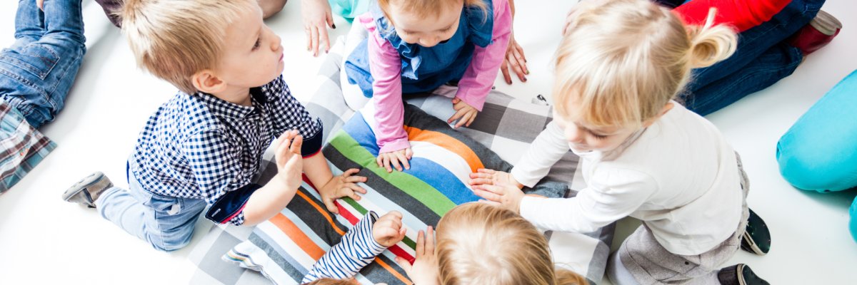 Eine Gruppe kleiner Kinder sitzt in einem Kreis auf einer bunt gestreiften Decke. Im Hintergrund sind Teile von Erwachsenen zu sehen.
