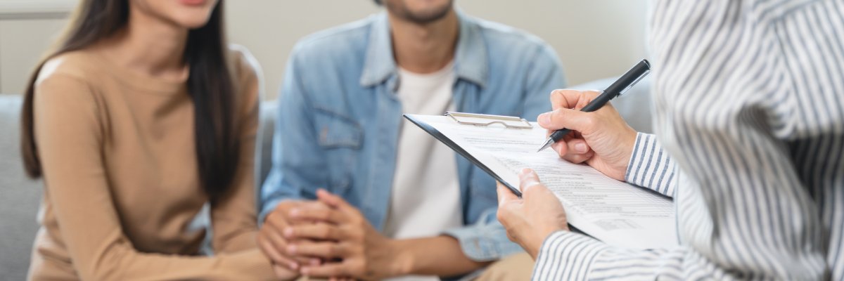 Eine Person in einem gestreiften Hemd schreibt auf einem Klemmbrett, während im Hintergrund ein lächelndes Paar sitzt und Händchen hält.