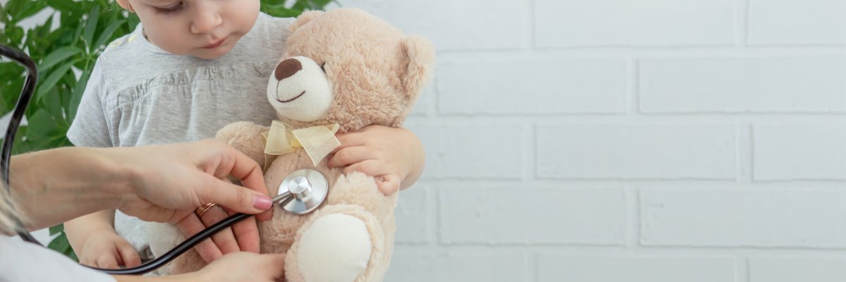 Eine erwachsene Person untersucht mit einem Stethoskop einen hellbraunen Teddybären, den ein kleines Kind hält.
