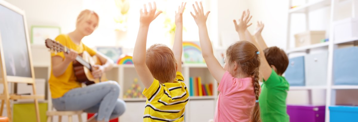 Eine Frau mit einer Gitarre und 3 Kinder die singen und die Arme hochheben