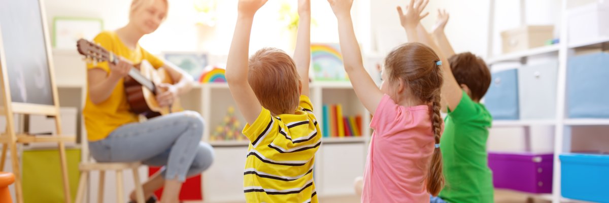 Eine Frau spielt Gitarre für drei Kinder, die auf einem Teppich sitzen und ihre Arme heben.