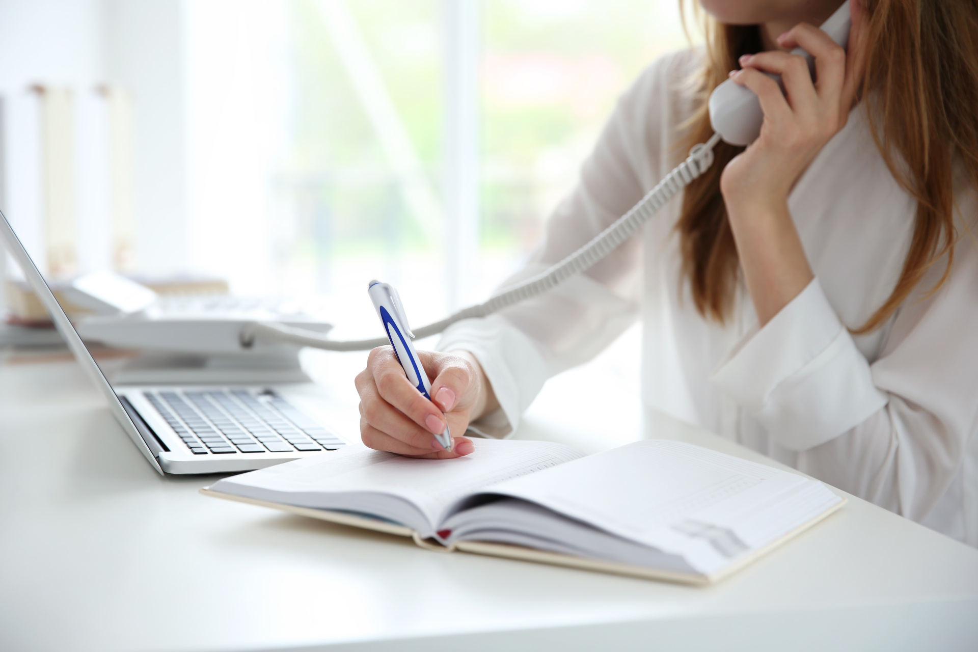 Person telefoniert und macht Notizen Eine Person in einem weißen Hemd telefoniert mit einem Festnetztelefon und schreibt gleichzeitig mit einem Stift in ein aufgeschlagenes Notizbuch.
