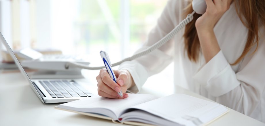 Person telefoniert und macht Notizen Eine Person in einem weißen Hemd telefoniert mit einem Festnetztelefon und schreibt gleichzeitig mit einem Stift in ein aufgeschlagenes Notizbuch.