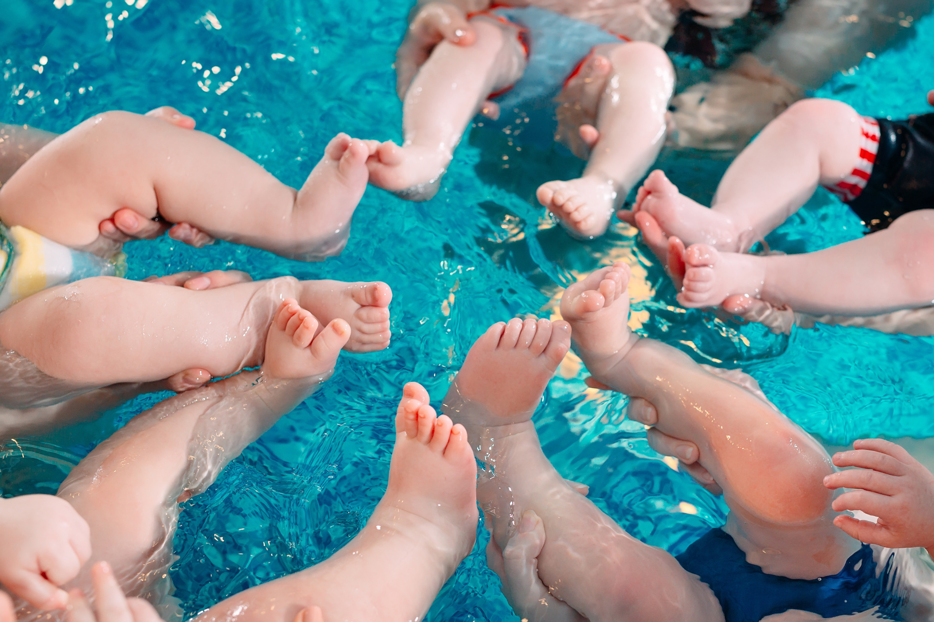 Babies im Schwimmbecken Vogelperspektive auf die Beine und Füße mehrerer Babys, die in einem hellblauen, klaren Schwimmbecken treiben. Die Beine sind kreisförmig in der Mitte angeordnet und bewegen sich im Wasser.