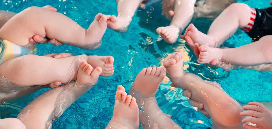 Babies im Schwimmbecken Vogelperspektive auf die Beine und Füße mehrerer Babys, die in einem hellblauen, klaren Schwimmbecken treiben. Die Beine sind kreisförmig in der Mitte angeordnet und bewegen sich im Wasser.