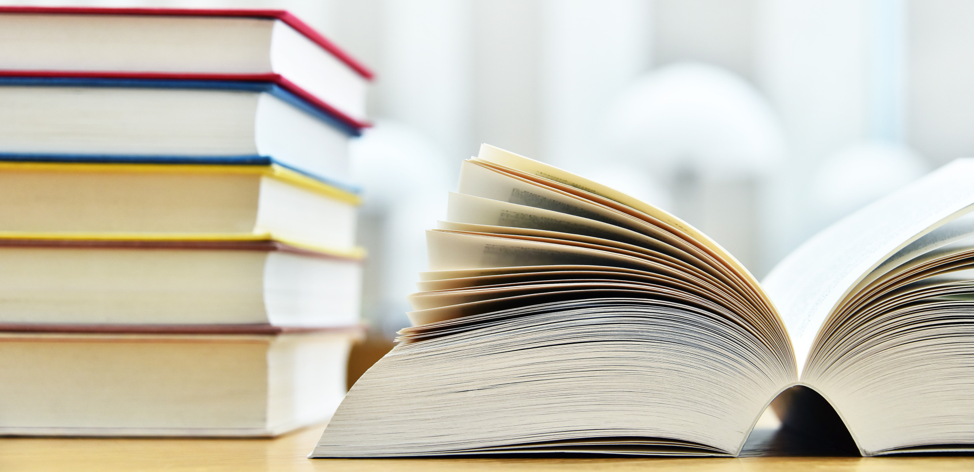Offenes Buch und Bücherstapel Ein aufgeschlagenes Buch liegt im Vordergrund, während im Hintergrund ein Stapel von Büchern mit farbigen Buchrücken zu sehen ist.