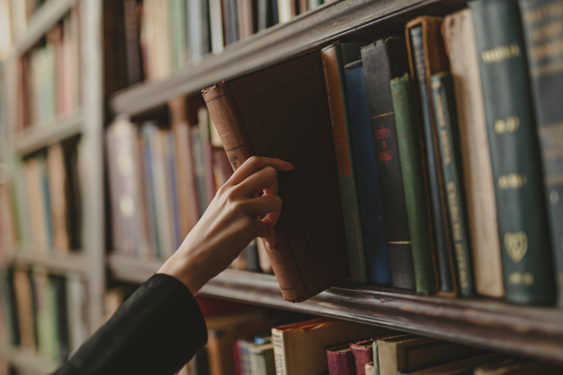 Buch aus dem Regal Eine Hand zieht ein braunes, gebundenes Buch aus einem hölzernen Bücherregal, in dem viele weitere alte Bücher stehen.