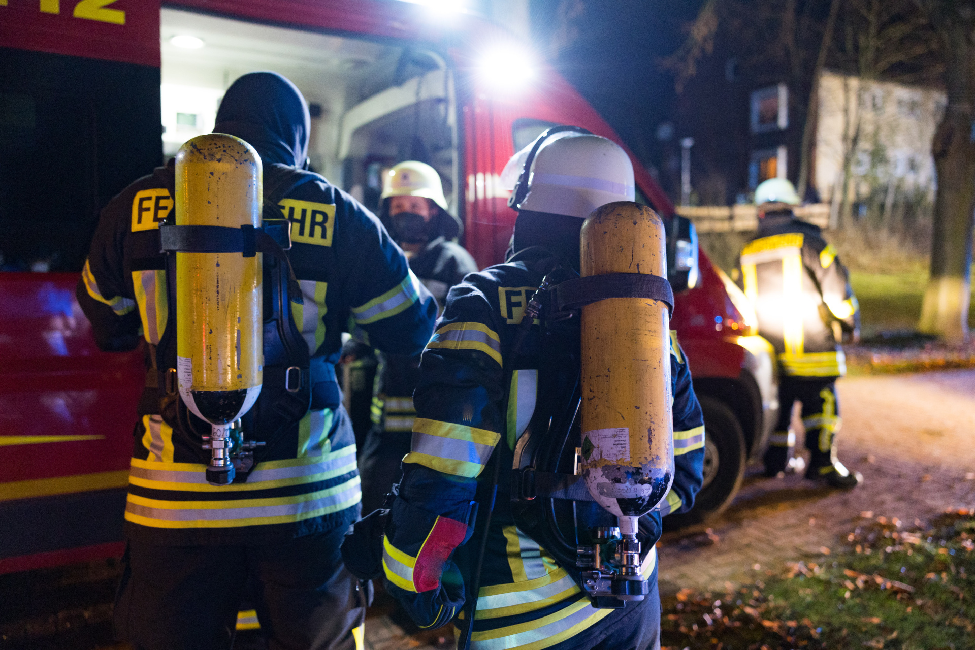 Mehrere Feuerwehrleute in dunkler Schutzkleidung mit gelben Reflexstreifen und Atemschutzflaschen auf dem Rücken stehen nachts vor einem roten Feuerwehrfahrzeug.