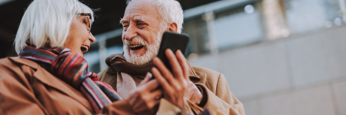 Ein lächelndes älteres Paar schaut gemeinsam auf ein Smartphone und trägt mehrere Einkaufstaschen.