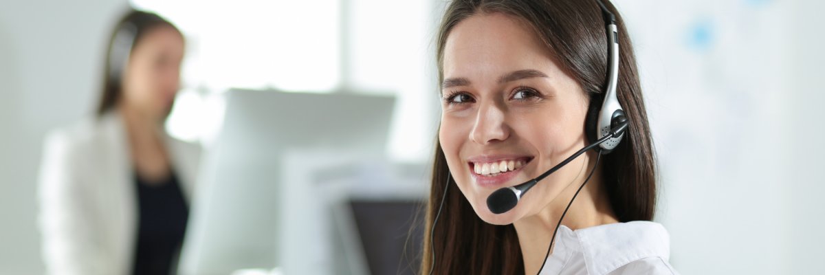 Mitarbeiterin mit Headset Eine lächelnde Frau trägt ein Headset und blickt in die Kamera. Im Hintergrund ist eine weitere Person mit Headset an einem Schreibtisch zu sehen.