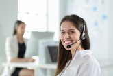 Eine lächelnde Frau trägt ein Headset und blickt in die Kamera. Im Hintergrund ist eine weitere Person mit Headset an einem Schreibtisch zu sehen.