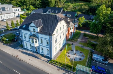 Luftaufnahme des Rathauses Overath. Davor liegt eine Grünfläche mit einem kreisförmigen Steinweg. Im Vordergrund ist eine Straße und daneben eine Parkfläche mit Autos zu sehen.