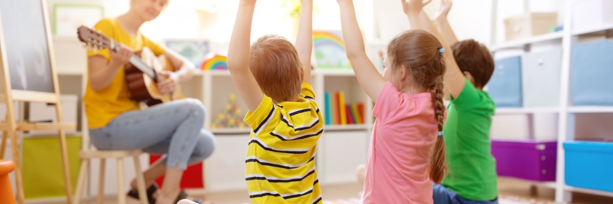 Eine Frau mit einer Gitarre und 3 Kinder die singen und die Arme hochheben