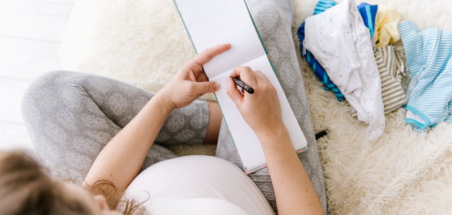 Eine Schwangere Frau sitzt auf dem Bett und schreibt eine Liste.