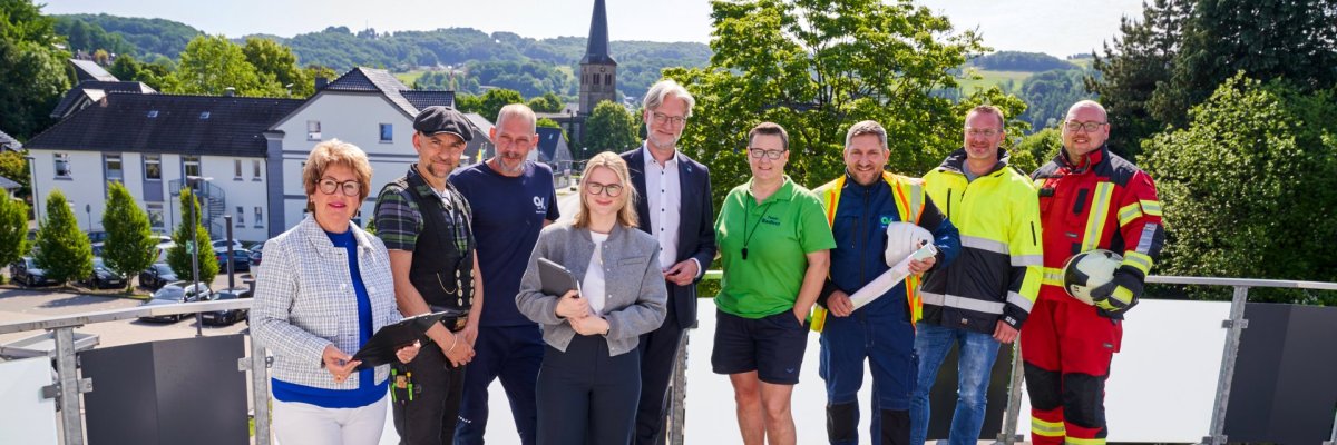 Mitarbeitende der Stadt Overath Acht Personen in unterschiedlicher Kleidung – darunter Business-Outfits, Arbeitskleidung und eine Feuerwehruniform – stehen nebeneinander auf einer grauen Terrasse.