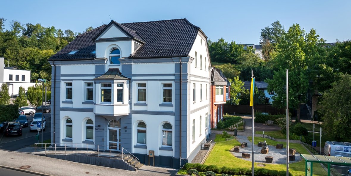Rathaus Overath Das weiße Gebäude des Rathauses mit grauen Akzenten und einem dunklen Ziegeldach. Über dem Eingang steht der Schriftzug „RATHAUS“.