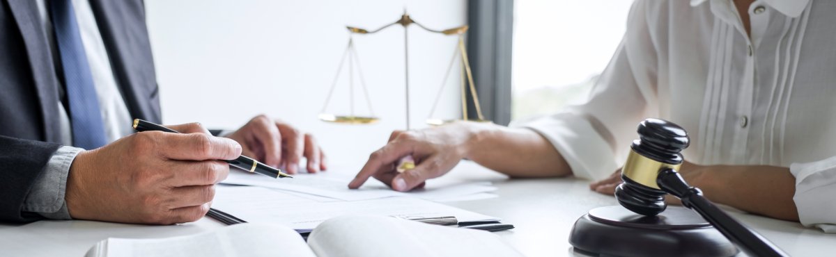 Zwei Personen die einen Gerichtstermin besprechen, es liegt ein geöffnetes Buch, eine Waage und ein Richterhammer auf dem Tisch