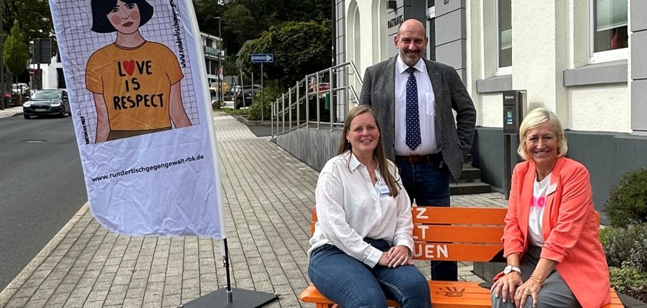 Auf einer orangenen Bank sitzen 2 Damen von Gleichstellung , der Bürgermeister steht hinter der Bank und neben der Bank steht ein Banner zum Thema Gewalt gegen Frauen