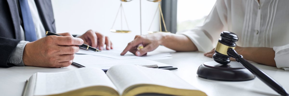 Zwei Personen die einen Gerichtstermin besprechen, es liegt ein geöffnetes Buch, eine Waage und ein Richterhammer auf dem Tisch